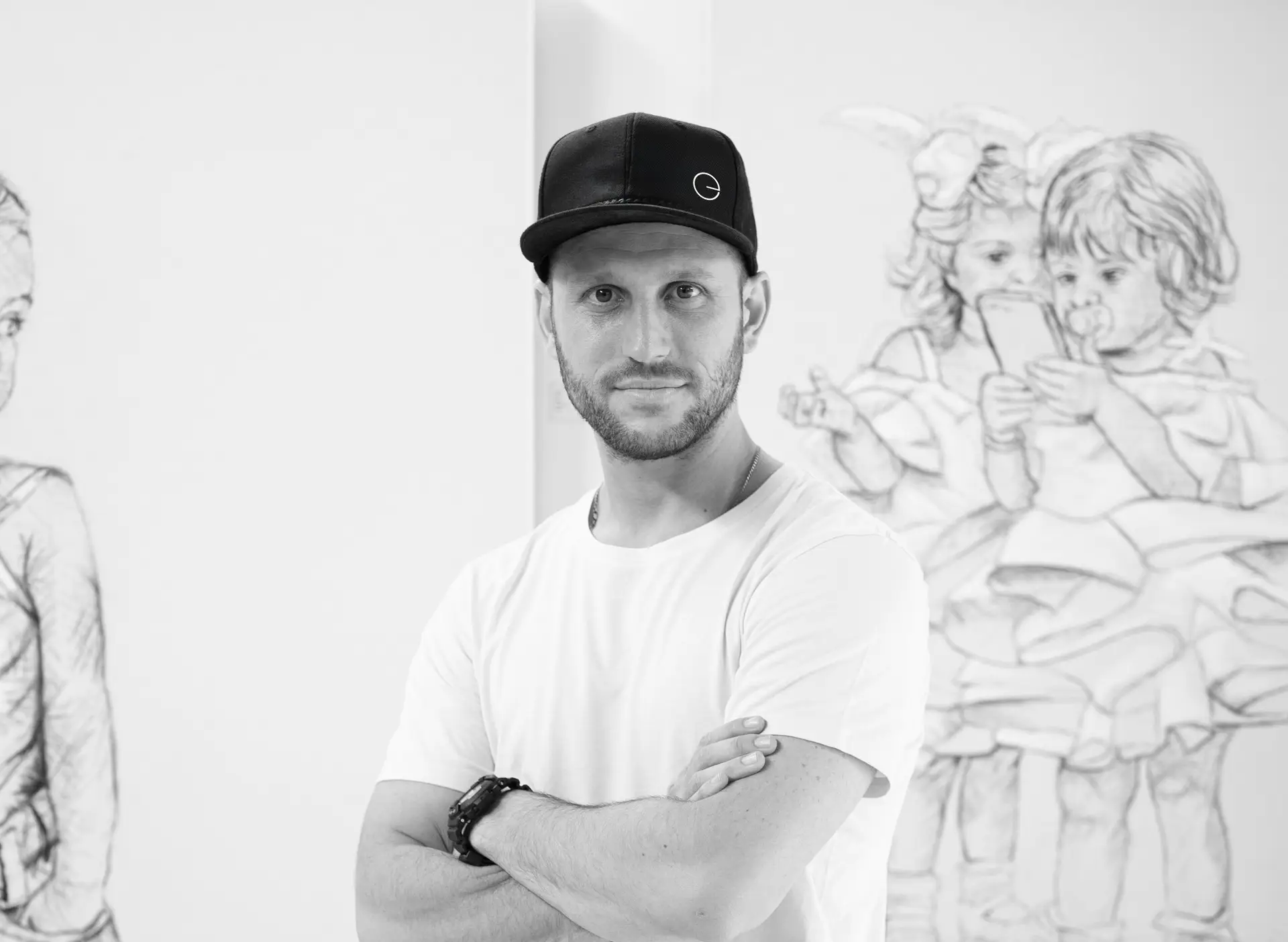 Man in a white t-shirt and black cap stands with his arms crossed in front of drawings of children. The artwork shows two kids holding a cellphone and another child to the left. The image is in black and white.